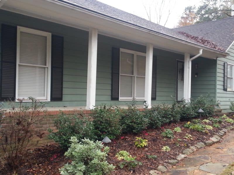 Lush landscaped front yard with shrubs and flowering plants in front of a green house, designed by John Russell Landscape Architect, showcasing professional outdoor landscaping.