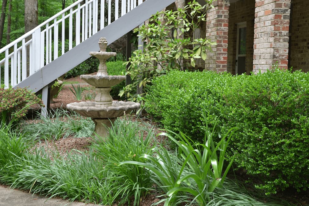 Stone garden fountain with lush greenery and brick house - John Russell Landscape Architect.
