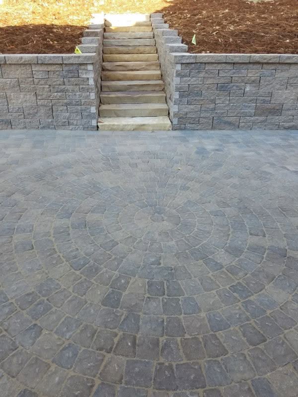 Natural stone patio with circular pattern and steps leading up to a landscaped area featuring stone retaining walls and soil preparation for planting.