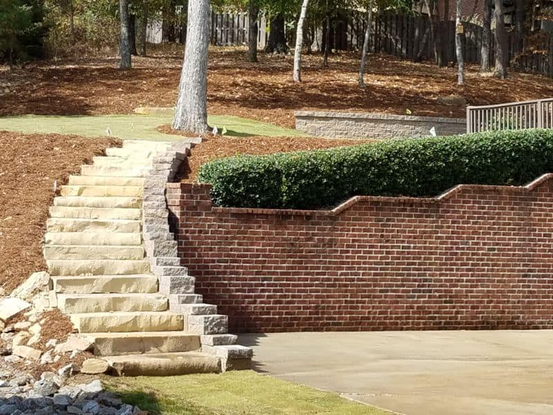 Natural stone staircase and red brick retaining wall in a landscaped garden with mature trees and shrubbery, designed by John Russell Landscape Architect, showcasing expert landscape architecture and hardscape craftsmanship.