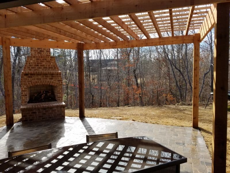 Rustic outdoor living space with brick fireplace, wooden pergola, and scenic yard view, designed by John Russell Landscape Architect for seamless indoor-outdoor entertaining.