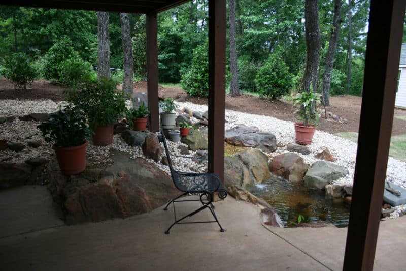 Rustic porch with landscaped garden featuring potted plants, rocks, and a small pond, created by John Russell Landscape Architect for outdoor living and backyard enhancement.
