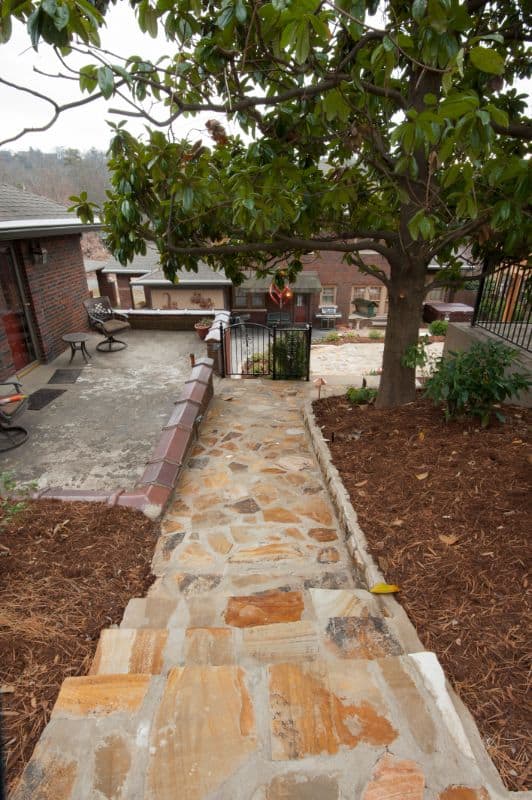 Natural stone pathway leading to a landscaped backyard featuring a large tree and manicured garden beds, designed by expert landscape architect John Russell.