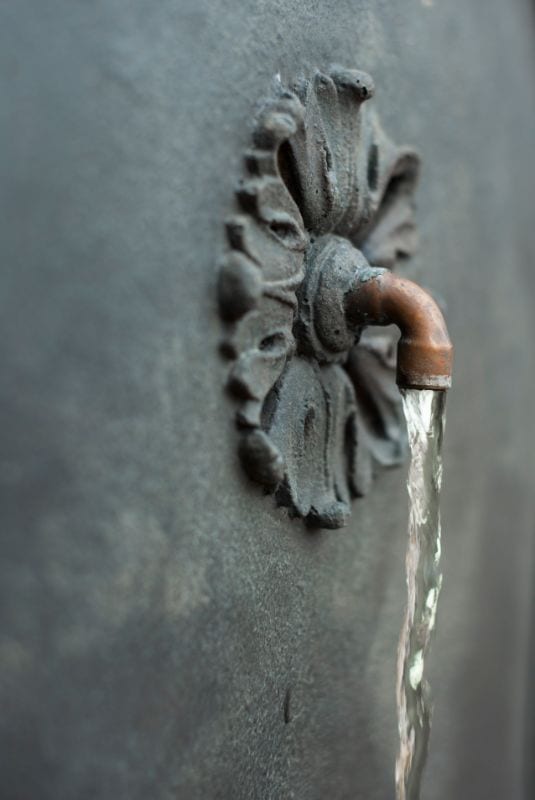 Rustic outdoor fountain with vintage metal spout on a textured wall, emphasizing garden water features and landscape design.