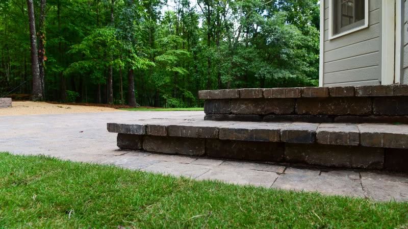 Natural stone patio with layered brick steps in a landscaped yard, designed by John Russell Landscape Architect, blending outdoor living space with surrounding greenery and trees.