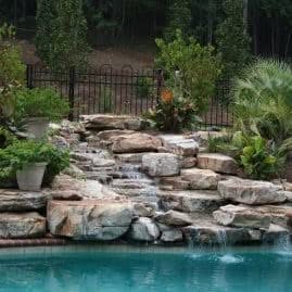 Rock waterfall landscape design with pool, water feature, lush greenery, tropical plants, garden landscaping, outdoor water fountain, natural stone waterfall, outdoor pool area, garden water feature, custom landscape architecture.