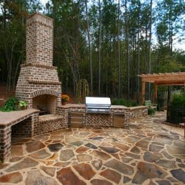 Fireplace outdoor brick patio in a backyard landscape design featuring a built-in brick fireplace, outdoor grill, and stone flooring, designed by John Russell Landscape Architect in a wooded setting.