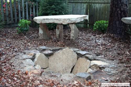 Rustic stone fire pit with natural rock seating area, surrounded by trees in a landscaped backyard, designed by John Russell Landscape Architect for outdoor entertainment and aesthetic appeal.