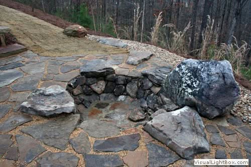 Natural stone fire pit with large rocks and flagstone patio, landscape design by John Russell Landscape Architect. Perfect for outdoor gatherings and backyard enhancements.