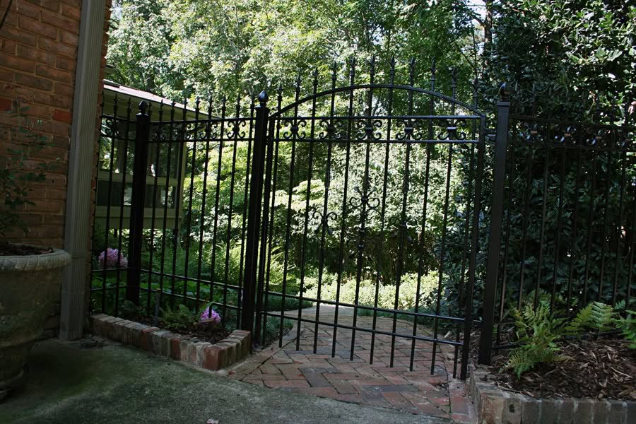 Iron garden gate with intricate scrollwork at landscape design by John Russell Landscape Architect, surrounded by lush greenery and brick pathway.
