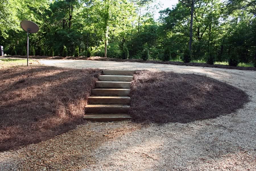 Raised landscape area with wooden steps and mulch, surrounded by lush green trees, designed by John Russell Landscape Architect for aesthetic curb appeal and functional outdoor space.