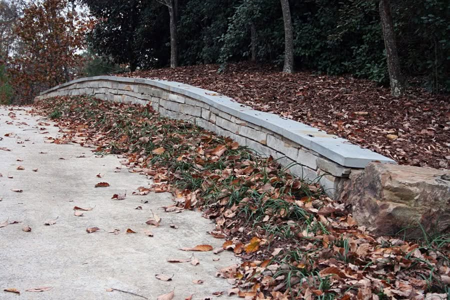 Natural stone retaining wall with fallen autumn leaves and lush landscape plants, designed by John Russell Landscape Architect, enhancing garden durability and aesthetic appeal.