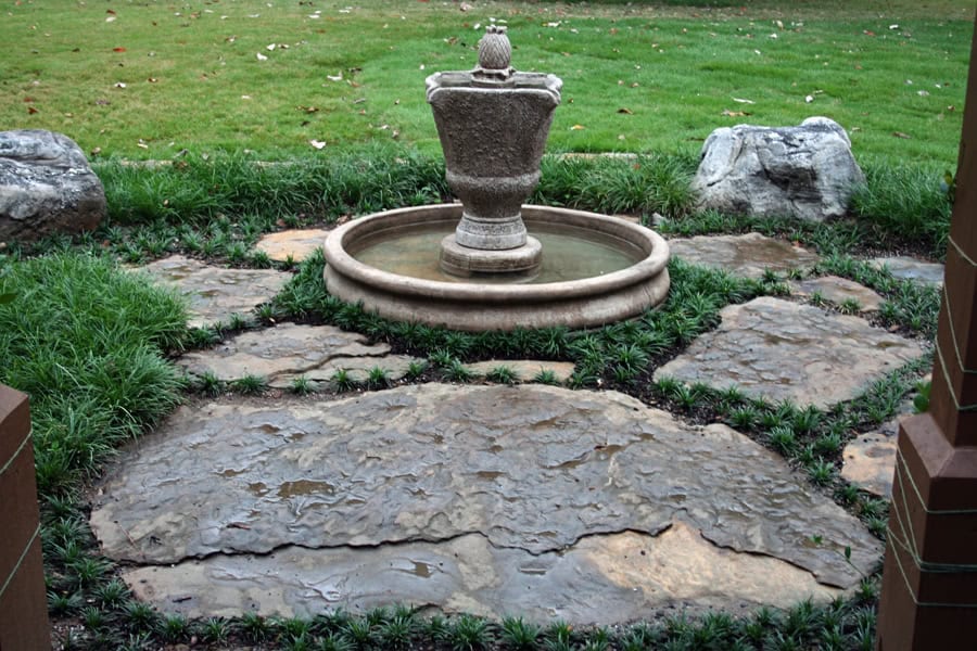 Elegant stone pathway with a decorative fountain feature, designed by John Russell Landscape Architect, creating a serene outdoor space with lush greenery and natural stone accents.