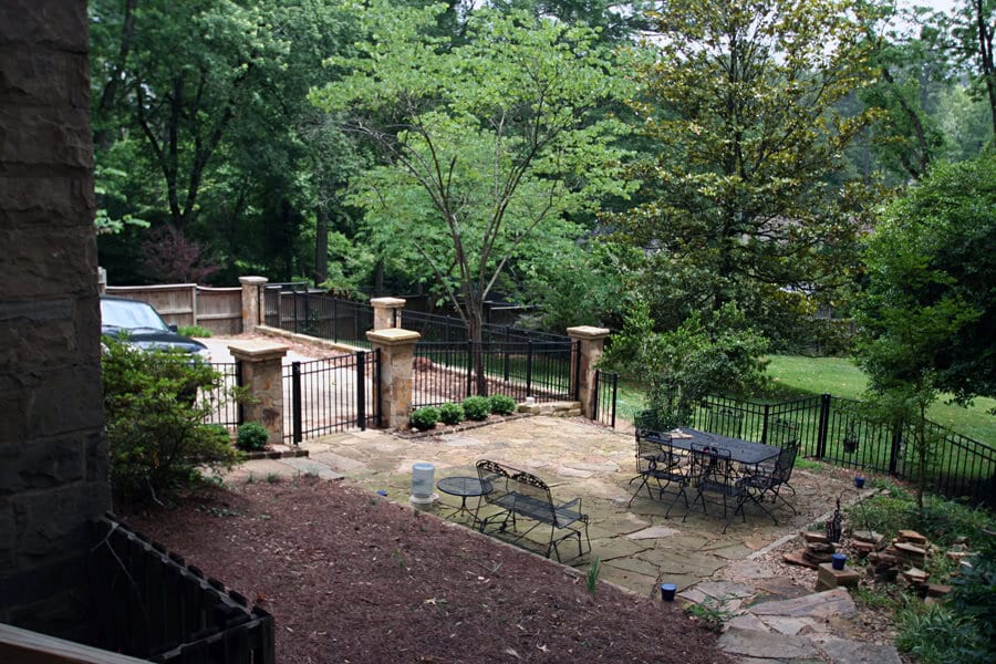 A scenic backyard patio with elegant wrought iron furniture, surrounded by lush green trees and a decorative black metal fence, designed by John Russell Landscape Architect.