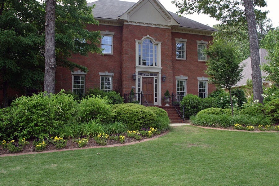 Lush landscaped front yard with colorful flowers, well-manicured lawn, and mature trees in front of a classic brick home.