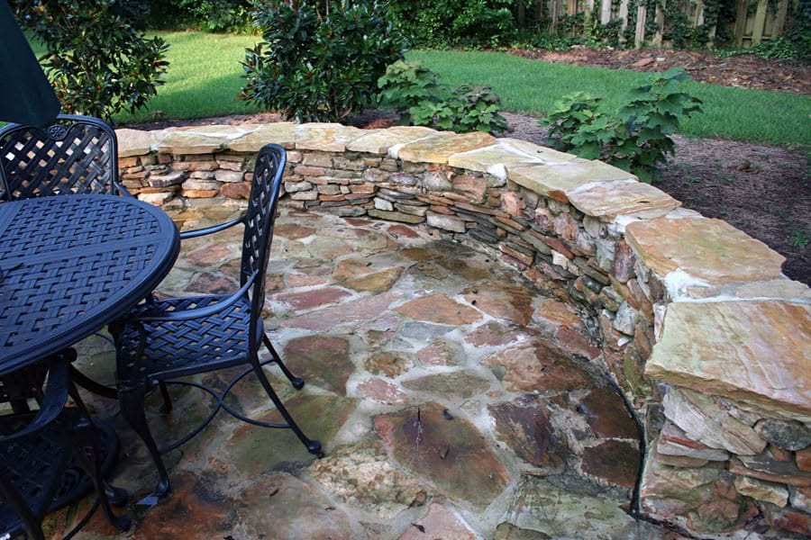 A beautiful stone outdoor patio with a curved stone planter and wrought iron furniture, designed by John Russell Landscape Architect. Perfect for relaxing and entertaining in a lush garden setting.