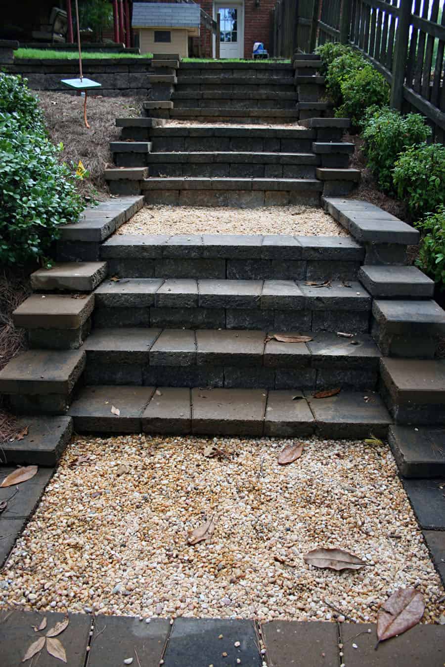Concrete paver outdoor staircase with black and gray stones, bordered by lush green shrubs, leading from backyard to upper patio with wooden railing, designed by John Russell Landscape Architect.