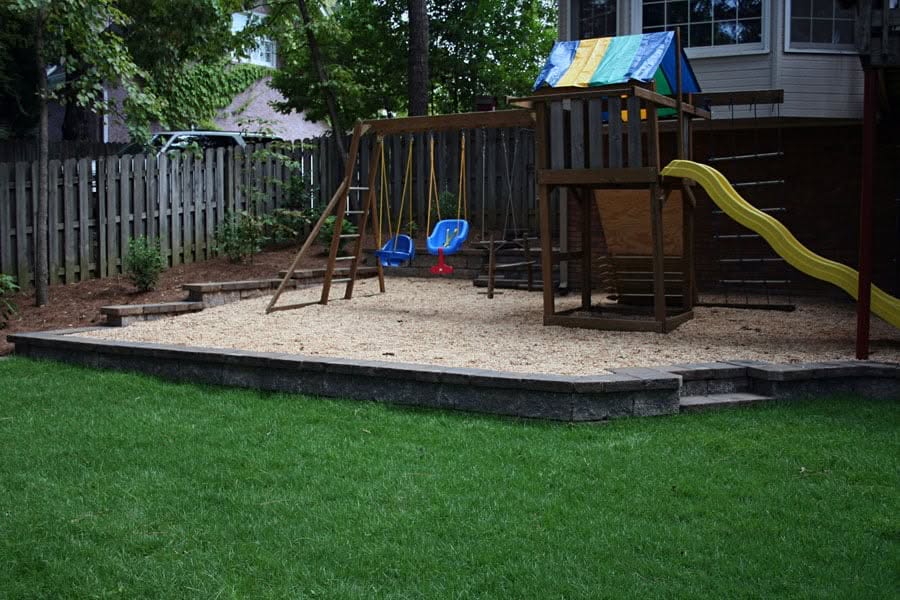 Colorful backyard playset with swings, slide, and sandbox area, surrounded by a wooden privacy fence and lush green lawn, designed by John Russell Landscape Architect for family outdoor enjoyment.