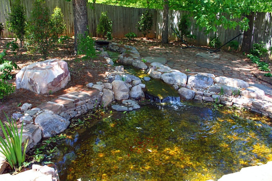 Rustic backyard pond with natural stone waterfall and lush greenery, designed by John Russell Landscape Architect. Ideal for enhancing outdoor spaces with water features and serene garden ambiance.