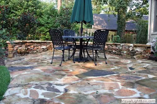 Natural stone patio with outdoor dining table and chairs, lush garden backdrop, scenic backyard landscape design.