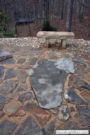 Handcrafted natural stone patio with flagstone pathway and earthy landscape design in a wooded backyard setting.