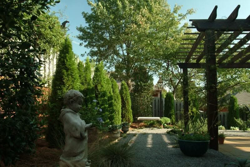 Lush backyard landscape design featuring mature trees, decorative sculptures, and a wooden pergola, created by John Russell Landscape Architect, showcasing expert outdoor space planning and lush greenery.